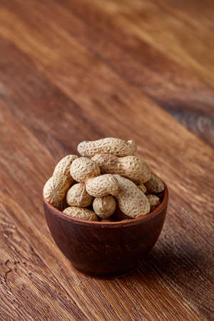 Unpeeled peanuts in wooden bowl over rustic wooden background closeup, selective focus. Some copy...