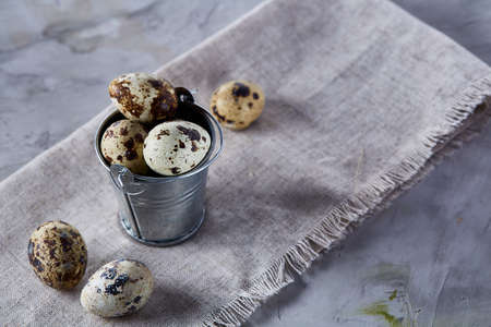 Small metal decorative bucket filled with fresh quail eggs on homespun napkin over white textured...