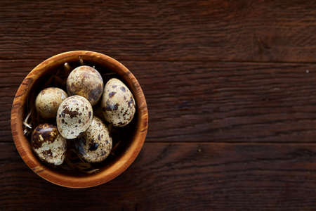 Fresh quail eggs in a wooden bowl on a dark wooden background, top view, close-up. Some copy space for your inscription. Textured background emphesize the still life. Healthy lifestyle concept.の写真素材