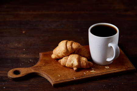 Breakfast with croissants and black coffee on wooden cutting board over rustic background, close-up, selective focusの写真素材