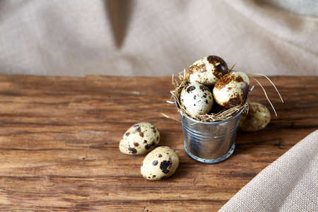 Small metal decorative bucket filled with fresh quail eggs on wooden board over white textured...