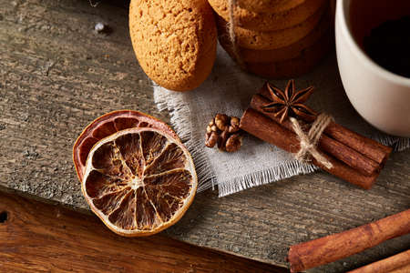 Traditional Christmas tea concept with a cup of hot tea, a large set of cookies on a homespun napkin, cinnamon sticks bunch, a couple of dried oranges and a branch of a spruce tree on a wooden table, selective focus. Festive concept.の写真素材