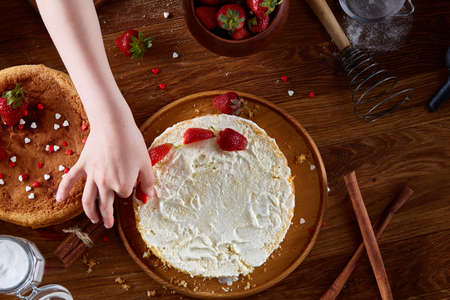 Top view of girl's hands putting strawberries on cake, close-up, selctive focus. Cooking dessert in...