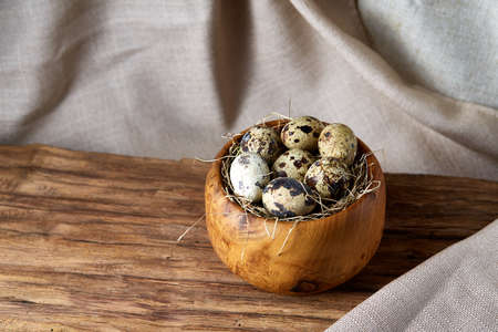 Fresh spotted quail eggs in a wooden bowl on a homespun tablecloth, top view, close-up, blurred.