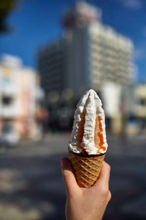 A first person view, girl walking along the road, holding ice cream in her hands and eating it in front of the modern building background, shallow depth of field, vertical. Yummy vanilla ice-cream. Sweet refreshing summer food concept.の写真素材