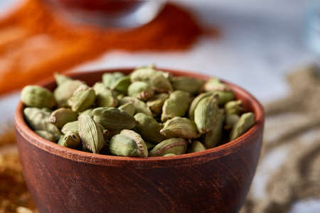 Green cardamon in ceramic bowl among ground scattered spicies on white textured background close-up,...