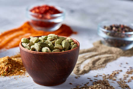 Green cardamon in ceramic bowl among ground scattered spicies on white textured background close-up,...