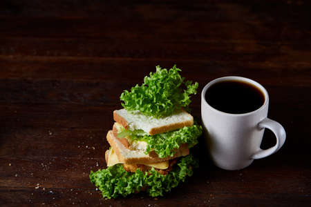 Breakfast table with fresh cheese and lettuce sandwich and black coffee in white ceramic mug on rustic wooden background, close-up, selective focus. Breakfast or lunch background. Tasty delectable appetizer. Food concept.の写真素材