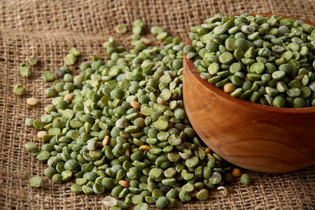 An overturned wooden bowl with organic green peas on burlap tablecloth, close-up, top view,...