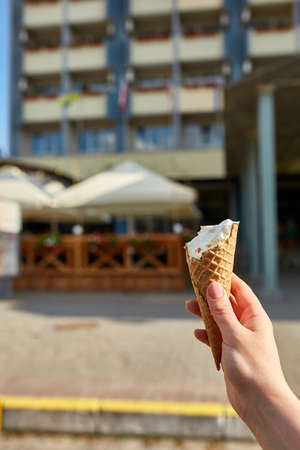 A first person view, a person walking along the road, holding ice cream in his hands and eating it on the retro car background, shallow depth of field. Delicious creamy dessert. Bitten vanilla cone. Sweet refreshing summer food concept.の写真素材