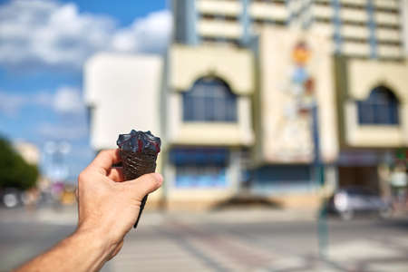 A first person view, a person walking along the road, holding ice cream in his hands and eating it on the casual city background, shallow depth of field, vertical. Bitten chocolate cone in tender woman's hand. Sweet refreshing summer food concept.の写真素材
