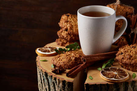 Christmas concept with a cup of hot tea, a large set of cookies, cinnamon sticks bunch, a couple of dried oranges, anise star and a branch of a spruce tree on a round log over rustic wooden background, selective focus.の写真素材