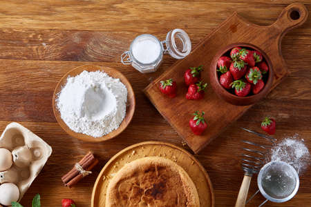 Delicious cake with fresh organic strawberries and kitchen utensils, top view, close-up, selctive focus. Sweet homemade dessert surrounded by natural ingredients on wooden table. Rustic background. Tasty morning pastry. Homemade food concept.の写真素材