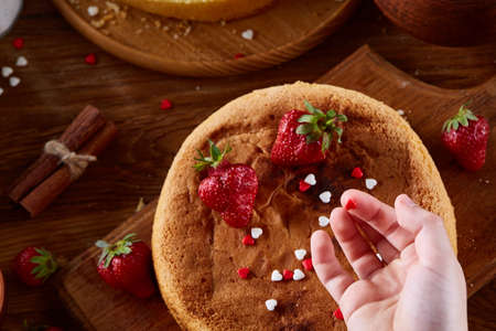 Close up of girl's hands decorating top of delicious strawberry cake, close up, selective focus. Cooking dessert in the kitchen. Tasty pastry is on the wooden table surrounded with organic ingredients. Homemade food concept. Baking with pleasure.の写真素材