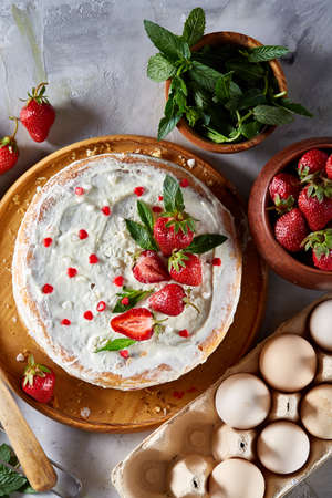 Strawberry tart covered with whipped sour cream surrounded by organic ingredients over white background, flat lay, selective focus. Homemade bakery with fresh eggs, mint leaves, organic strawberry and flour. Food concept. Bakery background.の写真素材