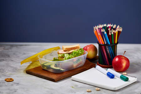 Concept of school lunch break with healthy lunch box full of sandwiches, banana, apples and school supplies on wooden desk, selective focus. There is a paper bag with some snack, emphesizing the concept. Time for study and relax.の写真素材