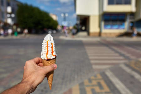 A first person view, a person walking along the road, holding ice cream in his hands and eating it in the quiet city background, shallow depth of field, vertical. Yummy chocolate dessert in strong man's hand. Sweet refreshing summer food concept.の写真素材