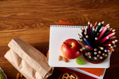 Tasty lunch with green lettuce and fresh vegetables and apple in transparent plastic container and different school supplies on wooden desk, selective focus, close-up. School lunch concept. Healthy eating concept.の写真素材