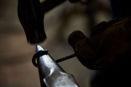Close-up of a blacksmith's hands manipulating a metal piece above his forge in the smithy, selective focus. Blacksmith striking a metallic object with his hammer. Blur pavement in background. Ancient manufacturing. Art and crafts concept.の写真素材