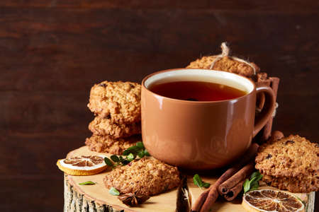 Christmas concept with a cup of hot tea, a large set of cookies, cinnamon sticks bunch, a couple of dried oranges, anise star and a branch of a spruce tree on a round log over rustic wooden background, selective focus.の写真素材