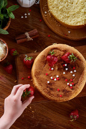 Close up of girl's hands decorating top of delicious strawberry cake, close up, selective focus. Cooking dessert in the kitchen. Tasty pastry is on the wooden table surrounded with organic ingredients. Homemade food concept. Baking with pleasure.の写真素材