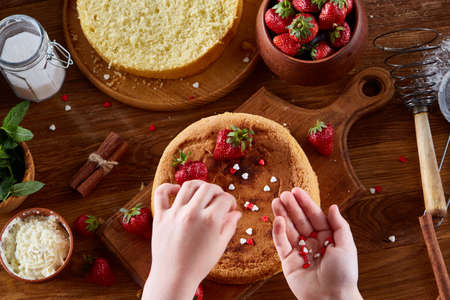 Close up of girl's hands decorating top of delicious strawberry cake, close up, selective focus. Cooking dessert in the kitchen. Tasty pastry is on the wooden table surrounded with organic ingredients. Homemade food concept. Baking with pleasure.の写真素材