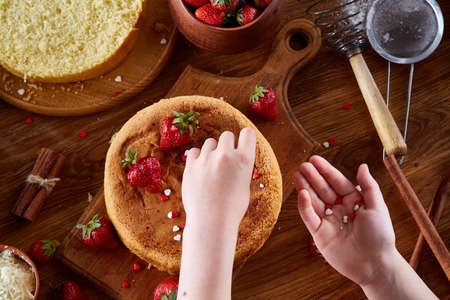 Close up of girl's hands decorating top of delicious strawberry cake, close up, selective focus. Cooking dessert in the kitchen. Tasty pastry is on the wooden table surrounded with organic ingredients. Homemade food concept. Baking with pleasure.の写真素材