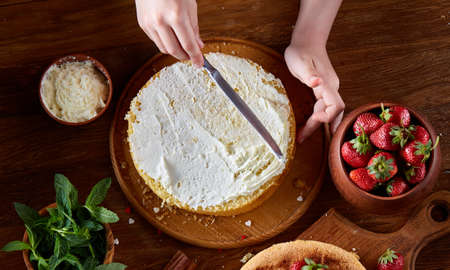 Close up of girl's hands adding some whipped sour cream on the top of delicious strawberry cake, selective focus. Tasty pastry is on the wooden table surrounded with organic ingredients. Homemade food concept. Baking with pleasure.の写真素材