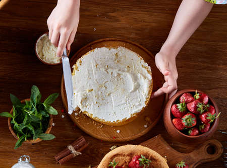 Close up of girl's hands adding some whipped sour cream on the top of delicious strawberry cake, selective focus. Tasty pastry is on the wooden table surrounded with organic ingredients. Homemade food concept. Baking with pleasure.の写真素材