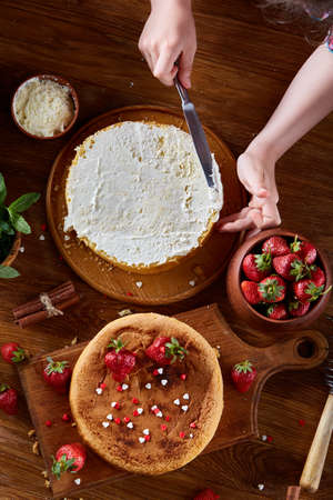 Close up of girl's hands adding some whipped sour cream on the top of delicious strawberry cake, selective focus. Tasty pastry is on the wooden table surrounded with organic ingredients. Homemade food concept. Baking with pleasure.の写真素材