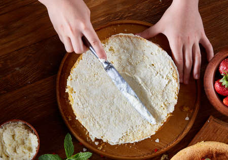 Close up of girl's hands adding some whipped sour cream on the top of delicious strawberry cake, selective focus. Tasty pastry is on the wooden table surrounded with organic ingredients. Homemade food concept. Baking with pleasure.の写真素材