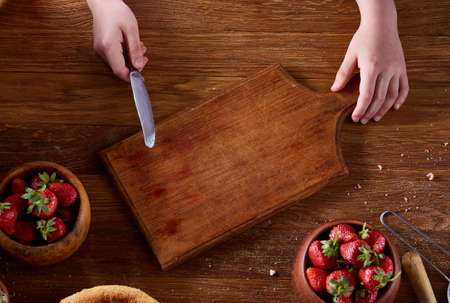 Knife in girl's hand cutting a fresh strawberry on wooden table, top view, selctive focus. Preparation for cooking something delicious. Natural organic berries. Nutritious vitamin. Healthy food concept.の写真素材