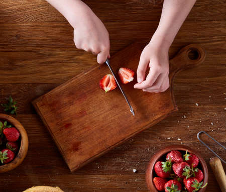 Knife in girl's hand cutting a fresh strawberry on wooden table, top view, selctive focus. Preparation for cooking something delicious. Natural organic berries. Nutritious vitamin. Healthy food concept.の写真素材