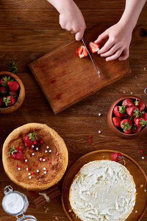 Knife in girl's hand cutting a fresh strawberry on wooden table, top view, selctive focus. Preparation for cooking something delicious. Natural organic berries. Nutritious vitamin. Healthy food concept.の写真素材