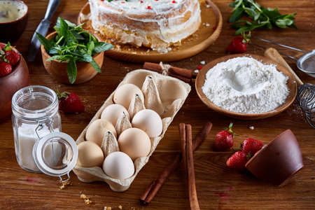 Strawberry tart covered with whipped sour cream surrounded by organic ingredients over wooden background, flat lay, selective focus. Homemade bakery with fresh eggs, mint leaves, organic strawberry and flour. Food concept. Bakery background.の写真素材