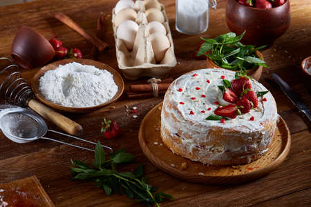 Strawberry tart covered with whipped sour cream surrounded by organic ingredients over wooden background, flat lay, selective focus. Homemade bakery with fresh eggs, mint leaves, organic strawberry and flour. Food concept. Bakery background.の写真素材