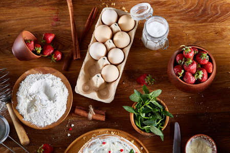 Strawberry tart covered with whipped sour cream surrounded by organic ingredients over wooden background, flat lay, selective focus. Homemade bakery with fresh eggs, mint leaves, organic strawberry and flour. Food concept. Bakery background.の写真素材