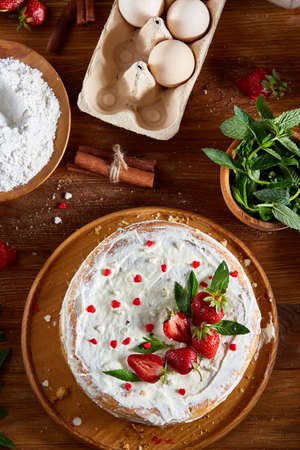 Strawberry tart covered with whipped sour cream surrounded by organic ingredients over wooden background, flat lay, selective focus. Homemade bakery with fresh eggs, mint leaves, organic strawberry and flour. Food concept. Bakery background.の写真素材