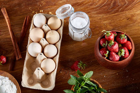 Strawberry tart covered with whipped sour cream surrounded by organic ingredients over wooden background, flat lay, selective focus. Homemade bakery with fresh eggs, mint leaves, organic strawberry and flour. Food concept. Bakery background.の写真素材