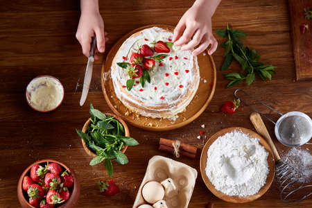Top view of girl's hands putting strawberries on cake, close-up, selctive focus. Cooking dessert in the kitchen. Tasty pastry is on the wooden table surrounded with organic ingredients. Homemade food concept. Baking with pleasure.の写真素材