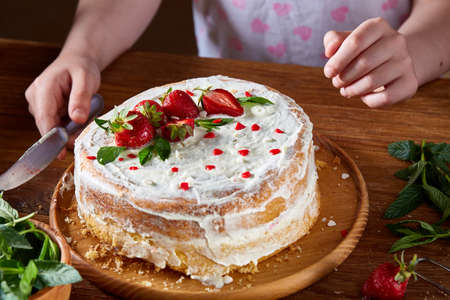 Top view of girl's hands putting strawberries on cake, close-up, selctive focus. Cooking dessert in the kitchen. Tasty pastry is on the wooden table surrounded with organic ingredients. Homemade food concept. Baking with pleasure.の写真素材