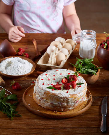 Top view of girl's hands putting strawberries on cake, close-up, selctive focus. Cooking dessert in the kitchen. Tasty pastry is on the wooden table surrounded with organic ingredients. Homemade food concept. Baking with pleasure.の写真素材