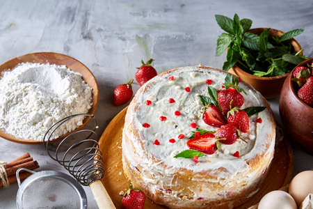 Strawberry tart covered with whipped sour cream surrounded by organic ingredients over white background, flat lay, selective focus. Homemade bakery with fresh eggs, mint leaves, organic strawberry and flour. Food concept. Bakery background.の写真素材
