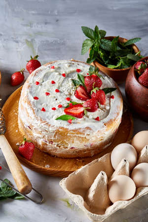 Strawberry tart covered with whipped sour cream surrounded by organic ingredients over white background, flat lay, selective focus. Homemade bakery with fresh eggs, mint leaves, organic strawberry and flour. Food concept. Bakery background.の写真素材