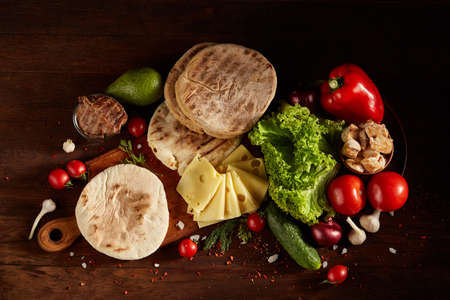 Plain pita on a cutting board with fresh vegetables and cheese on a wooden background, flat lay, selective focus, close-up. Still-life on a vintage background. Delicious barbecue background. Food concept.の写真素材