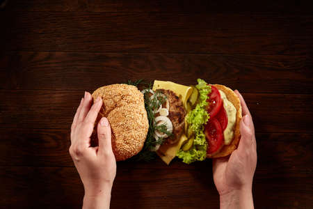 Woman's hands holding delicious fresh home made burger with piccoli, lettuce, cheese, onion, tomato, dill and beef on a dark wooden background, top view, close-up. Traditional American hamburger. Junk food full of calories. Fast food concept.の写真素材