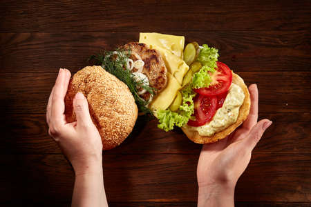 Woman's hands holding delicious fresh home made burger with piccoli, lettuce, cheese, onion, tomato, dill and beef on a dark wooden background, top view, close-up. Traditional American hamburger. Junk food full of calories. Fast food concept.の写真素材