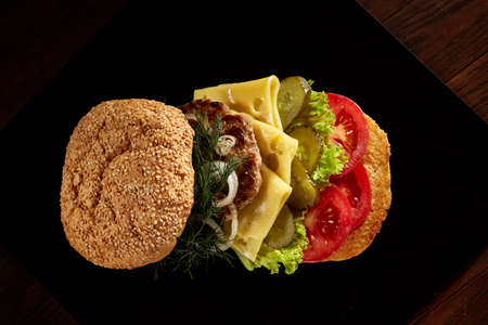 Big fresh burger arranged on black square plate on black wooden background, flat lay, close-up, selective focus. American classic burger menu. Artistic serving. Nutritious junk food. Fast food concept.の写真素材