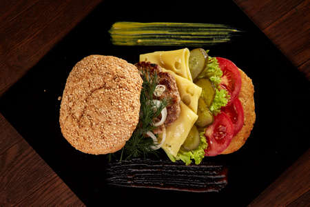 Big fresh burger with sauce arranged on black square plate on black wooden background, flat lay, close-up, selective focus. American classic burger menu. Artistic serving. Nutritious junk food. Fast food concept.の写真素材