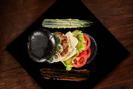 Black burger on square plate with spicy sauces on vintage wooden background, top view, close-up, selective focus. Artistic layout. Exotic hamburger stuffed with tomatoes, letucce, onion, piccoli and cheese. Nutritious junk food. Fast food concept.の写真素材